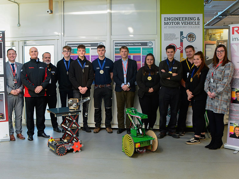A group of apprentices, College staff, including the Principal and employers standing behind two robots 