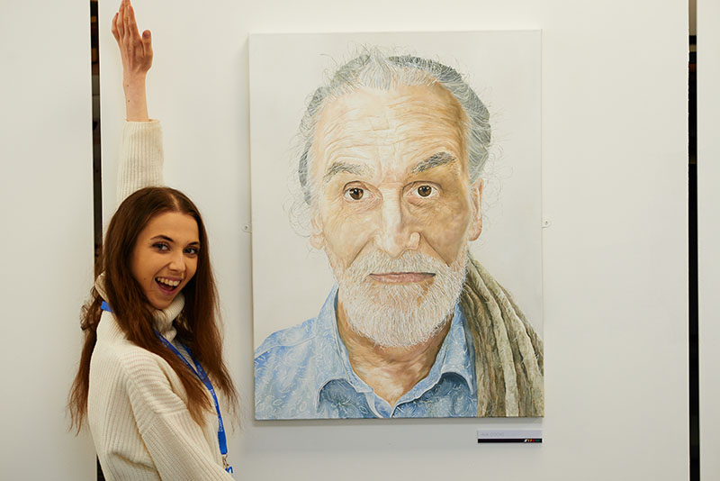 Female art student standing next to a large, lifelike portrait of a man with a beard.