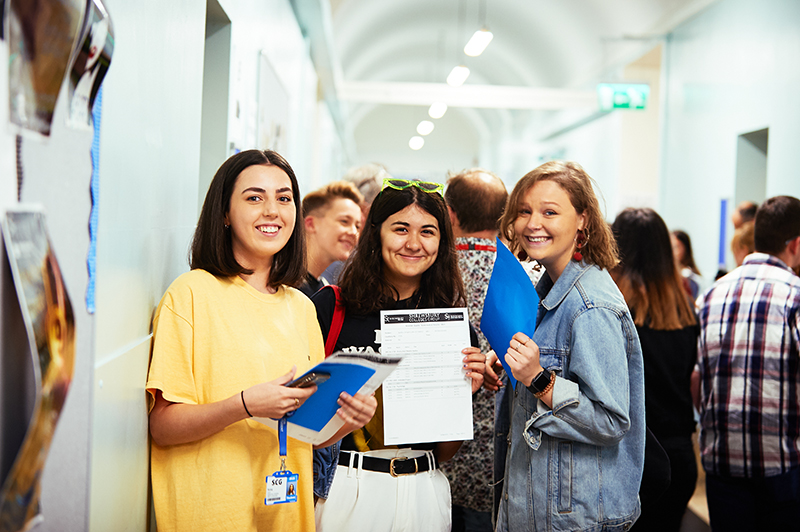 Three female students smiling with their exam results standing in front of a crowd of students.