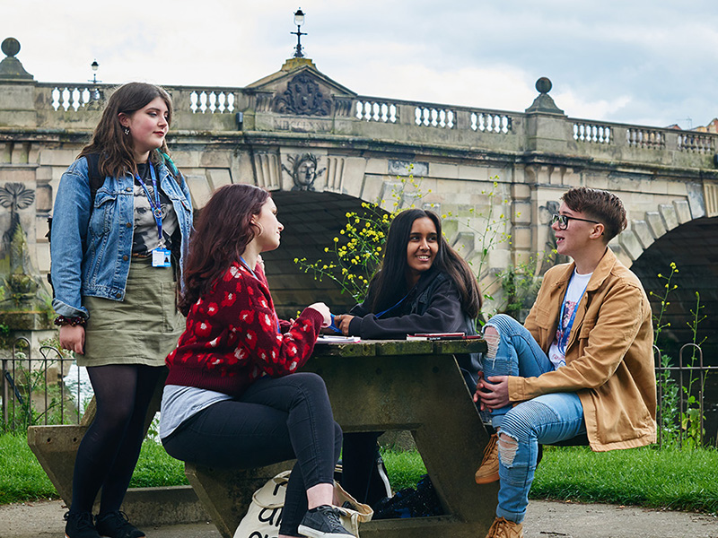 A group of students sat on a bench next to a river and bench in shrewsbury